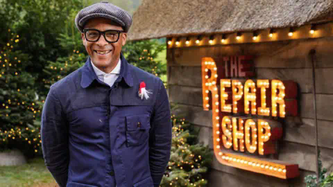 Jay Blades, wearing a grey cap, glasses and a buttoned up blue jacket, smiles at the camera with his hands behind his back. He is standing next to a brown shed, on which is a lit-up sign that says The Repair Shop.